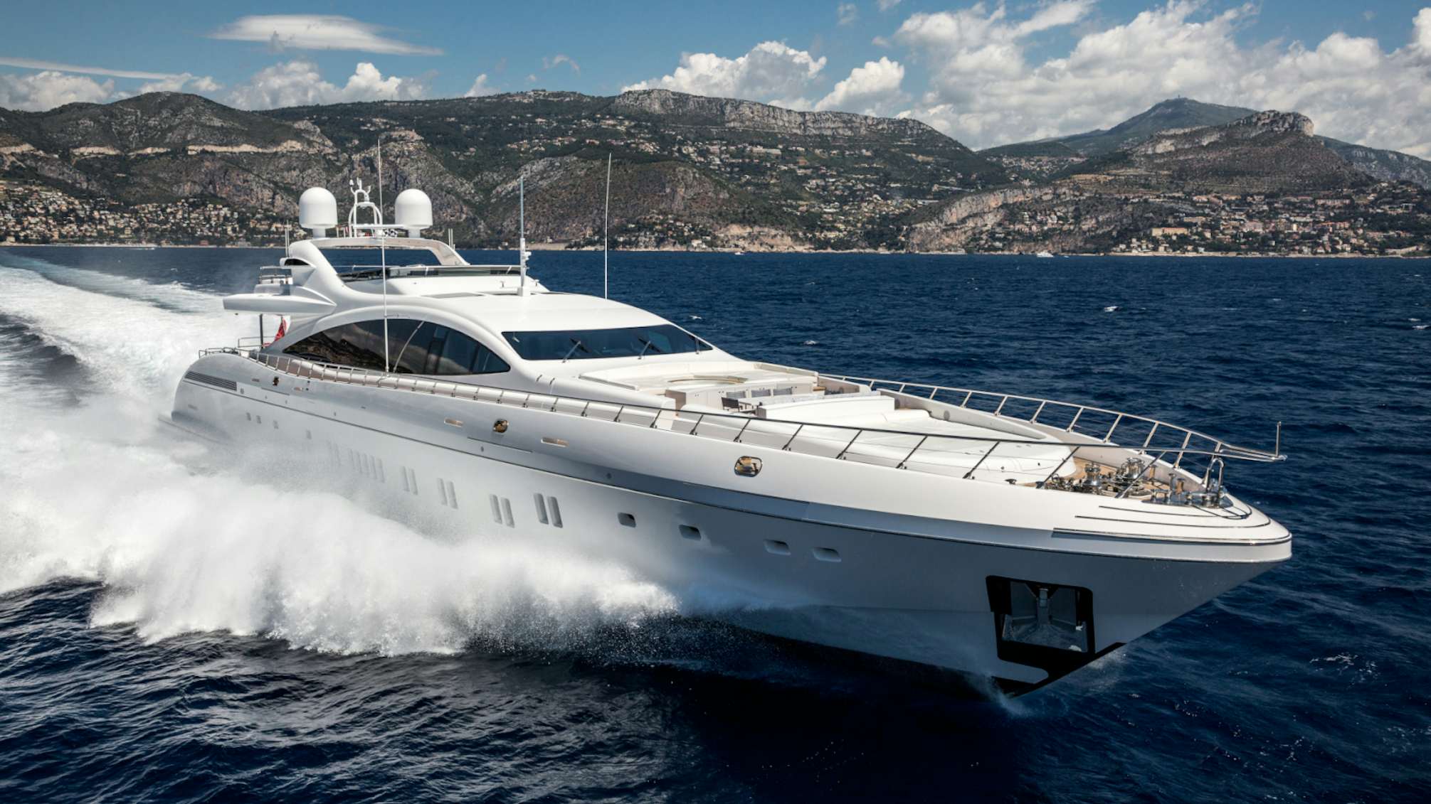 The Mangusta 165 yacht cutting through the blue waters, with a mountainous coastline in the background under a clear sky.