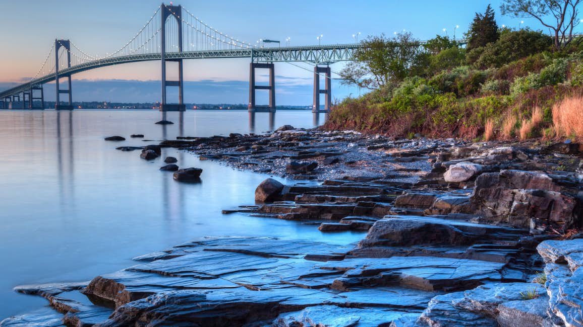 A captivating photo of the Newport Bridge and a rocky beach against a cloudy sunset backdrop during the Newport Charter Show.