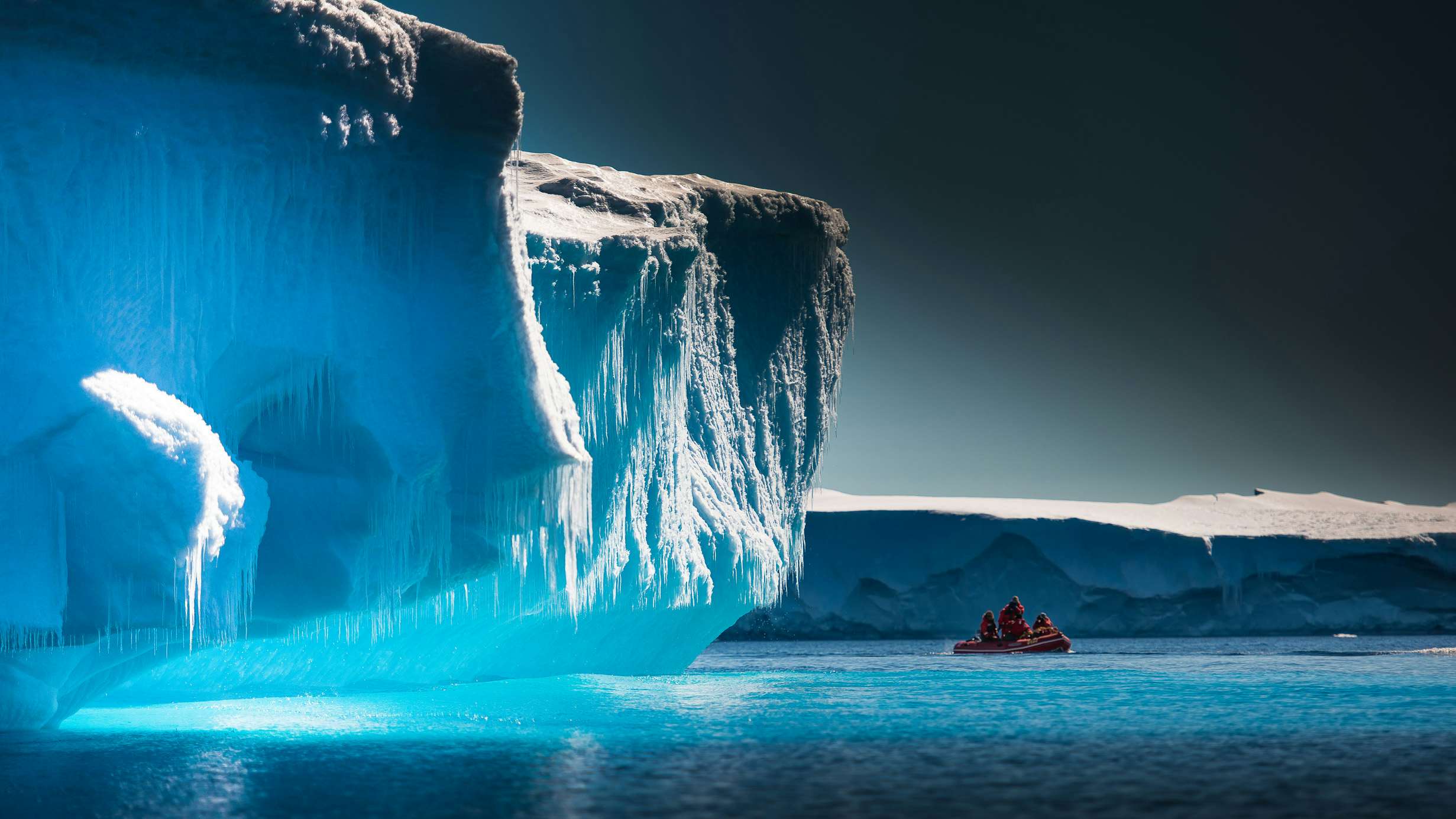 A small boat navigating the icy waters near a massive blue iceberg under a polar sky.