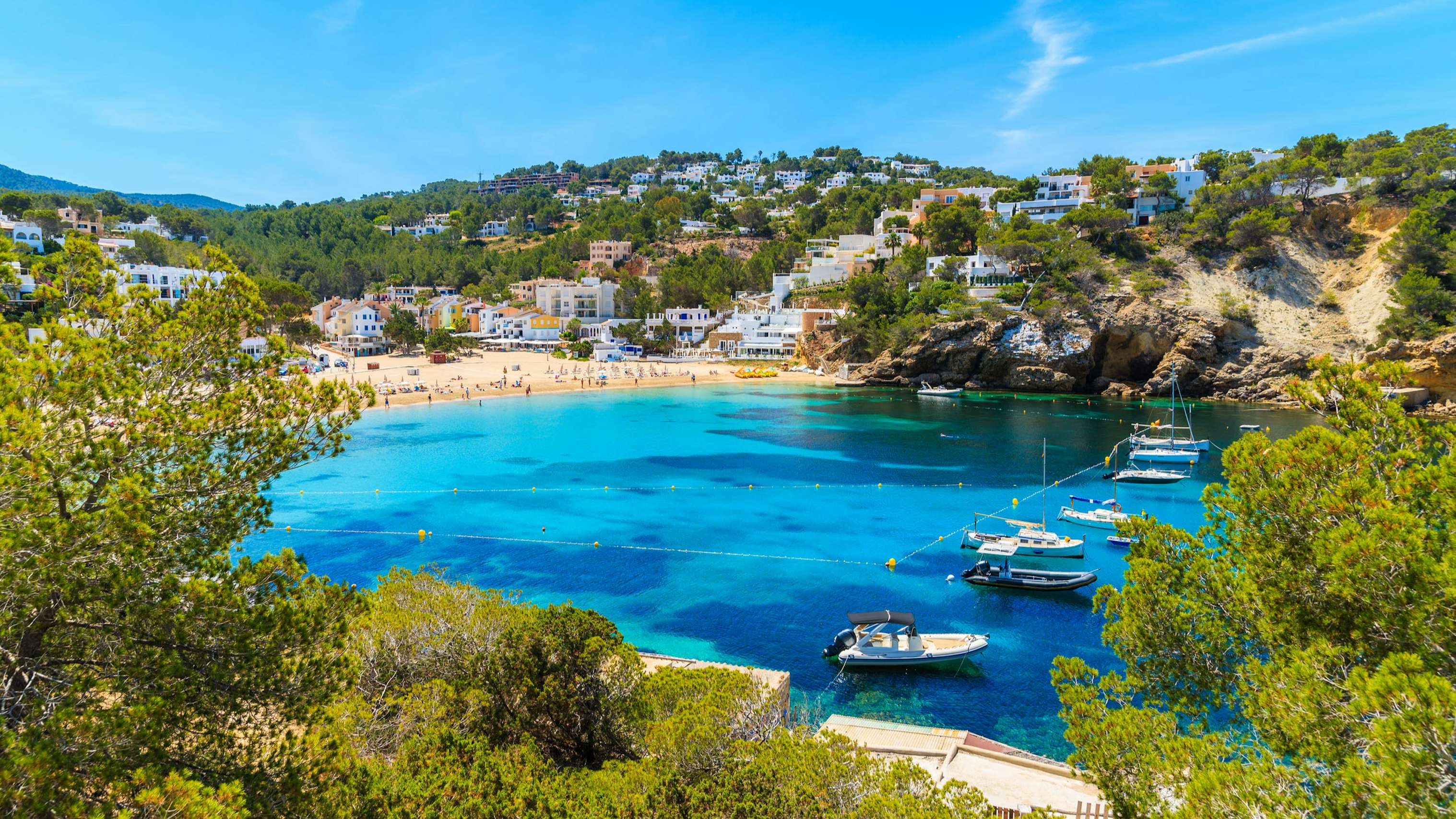 Yachts anchored in the tranquil azure waters of Cala Vadella, embraced by the lush green landscape.