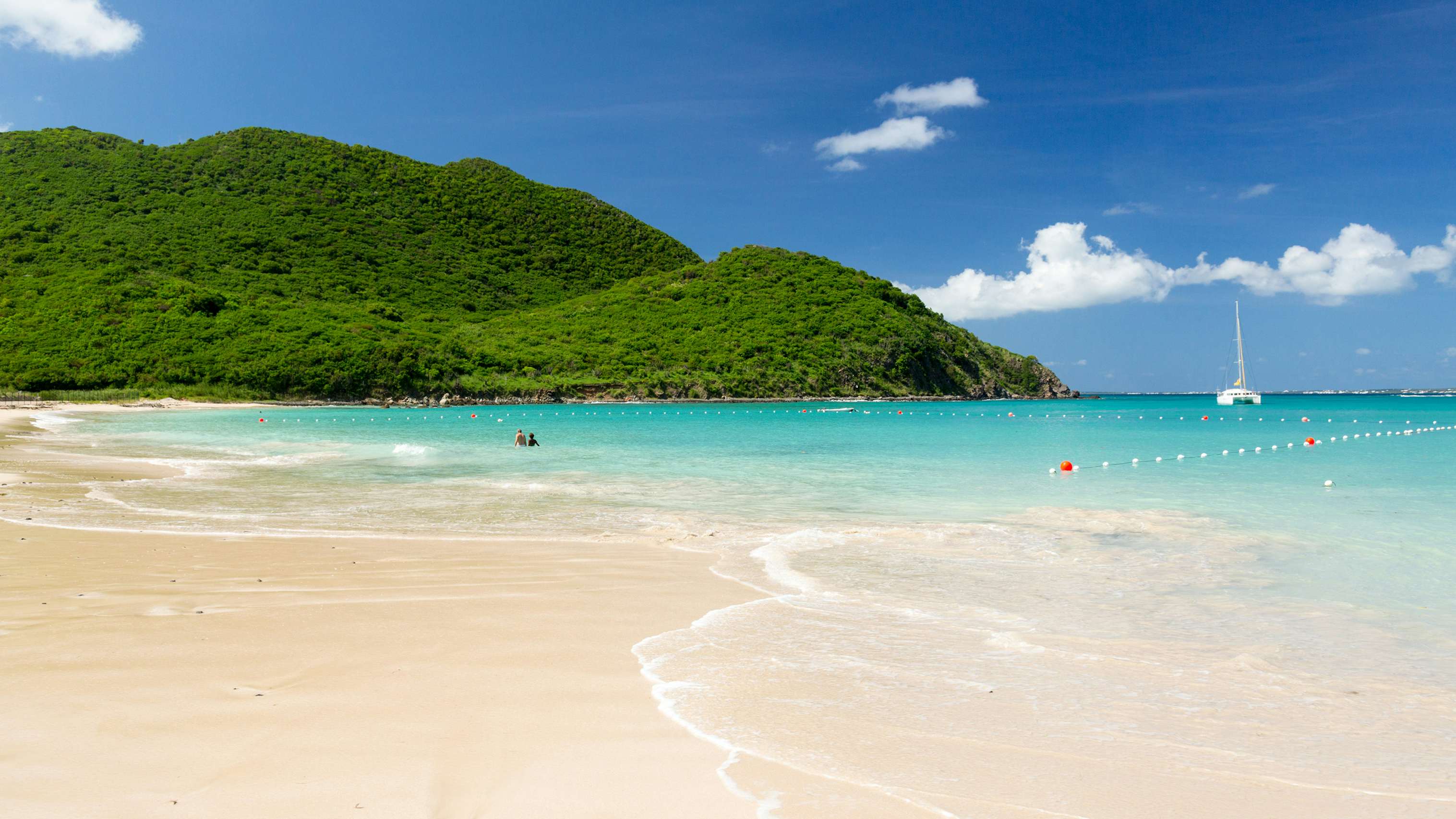 A picturesque scene of a sailboat gracefully cruising near a white sandy beach in St. Martin, inviting you to embark on the ultimate St. Maarten Yacht Charter experience.