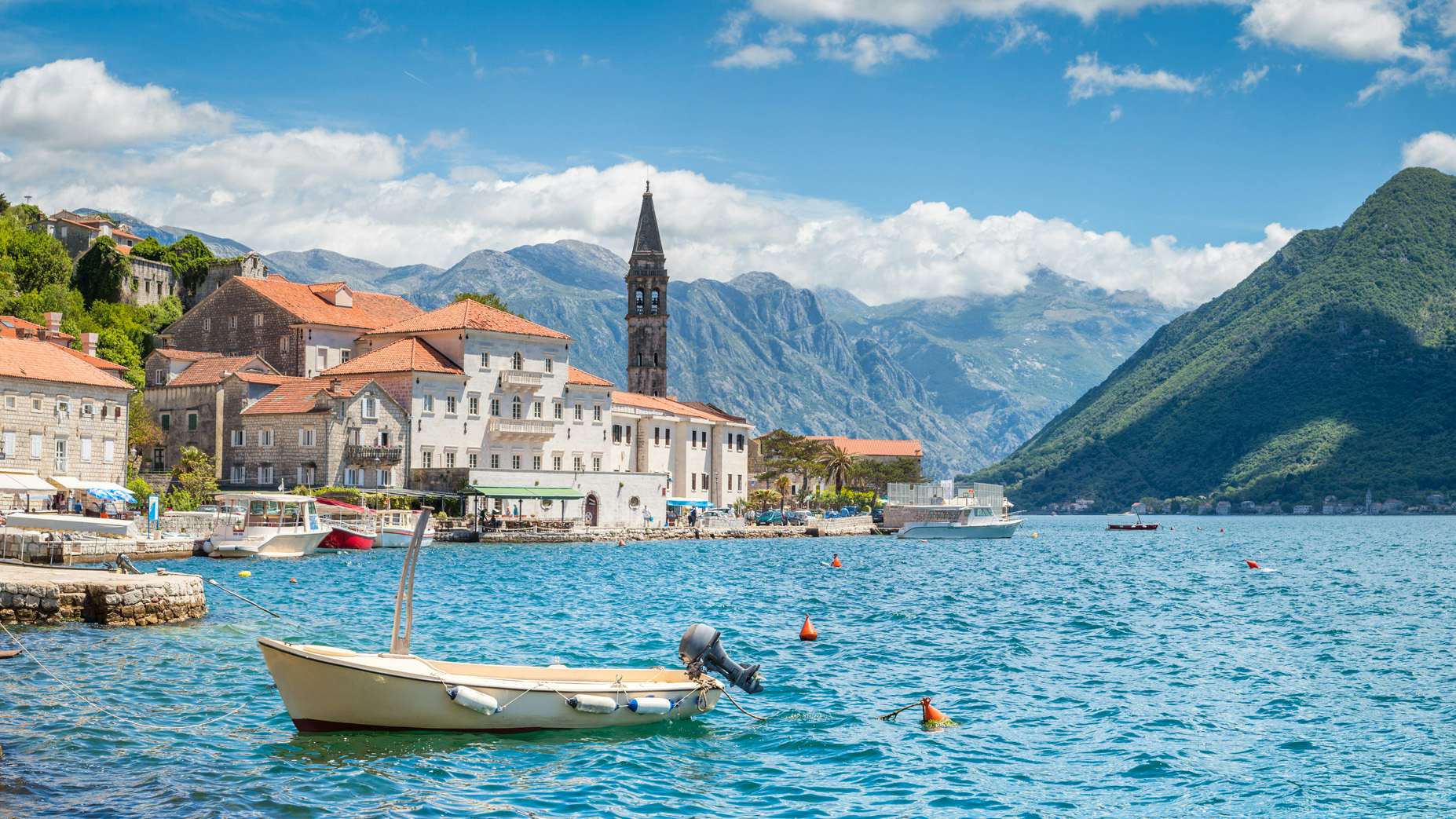 A small boat on the serene waters of Montenegro with a backdrop of mountains and historic buildings, ideal for yacht charter experiences by N&J.