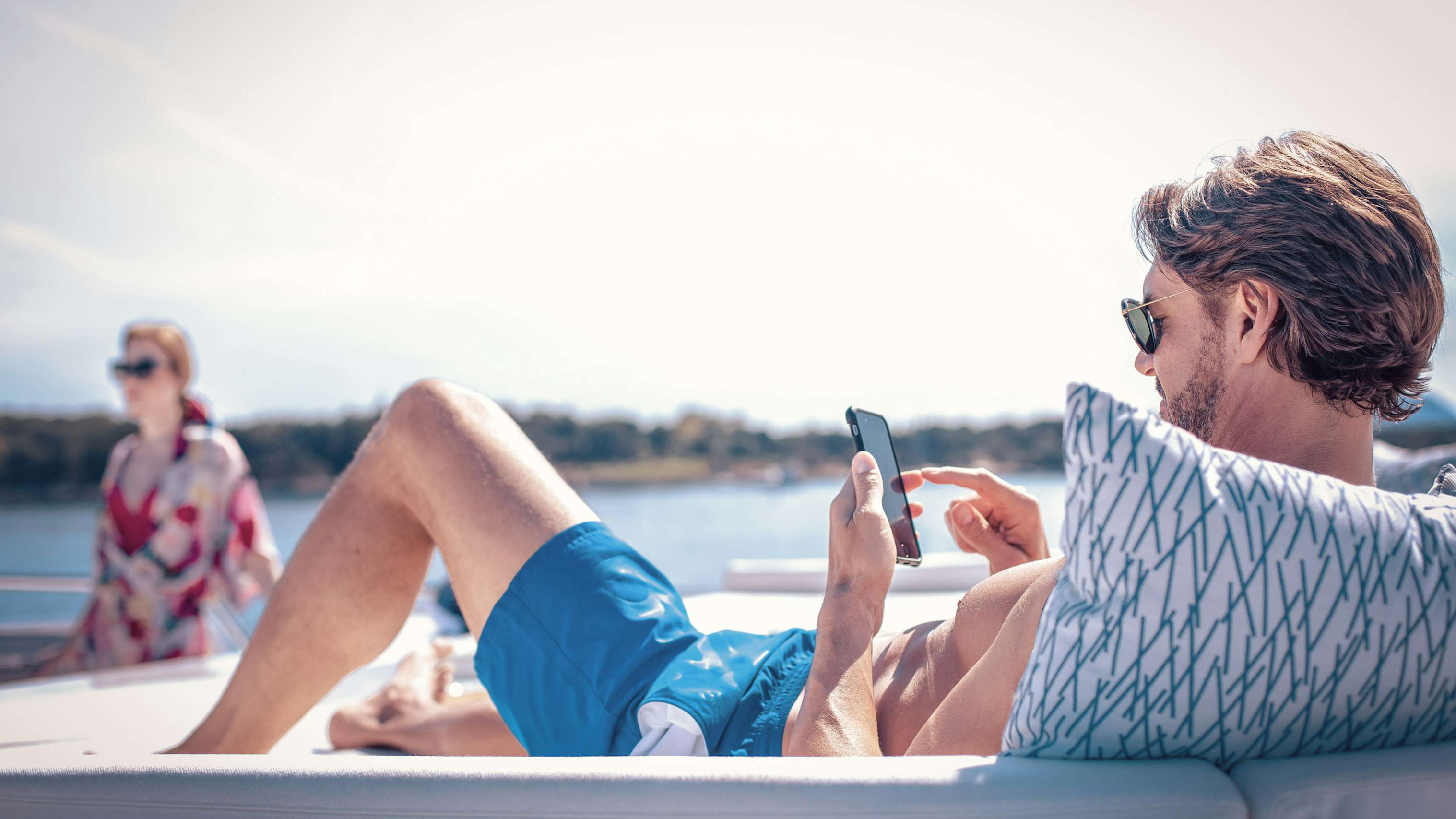 Man looking at mobile device while on sundeck with woman on background