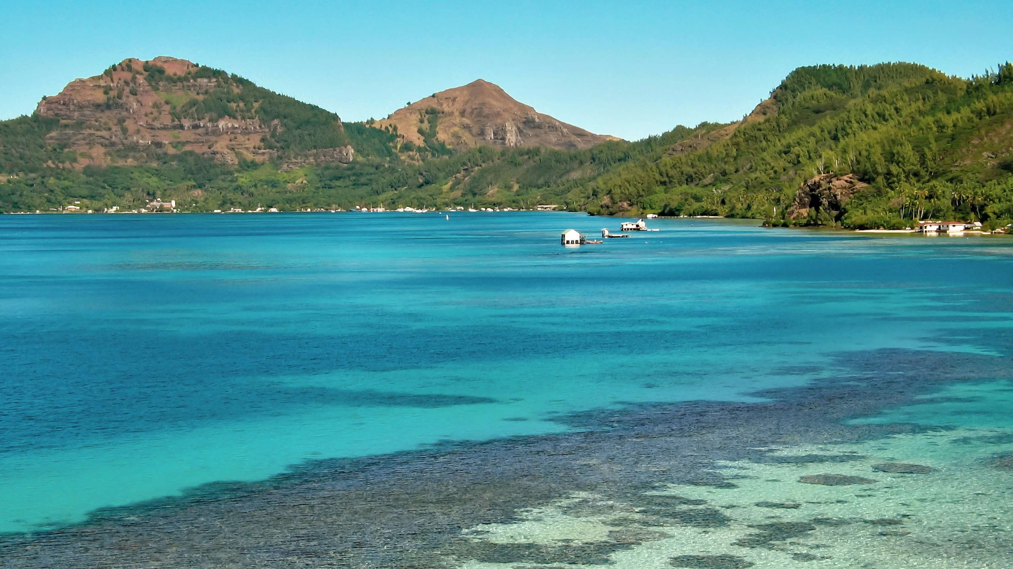 Tranquil coastal view of the Gambier Islands with clear blue waters and coral reefs in the foreground, anchored boats in the middle, and verdant hills in the background under a clear sky.