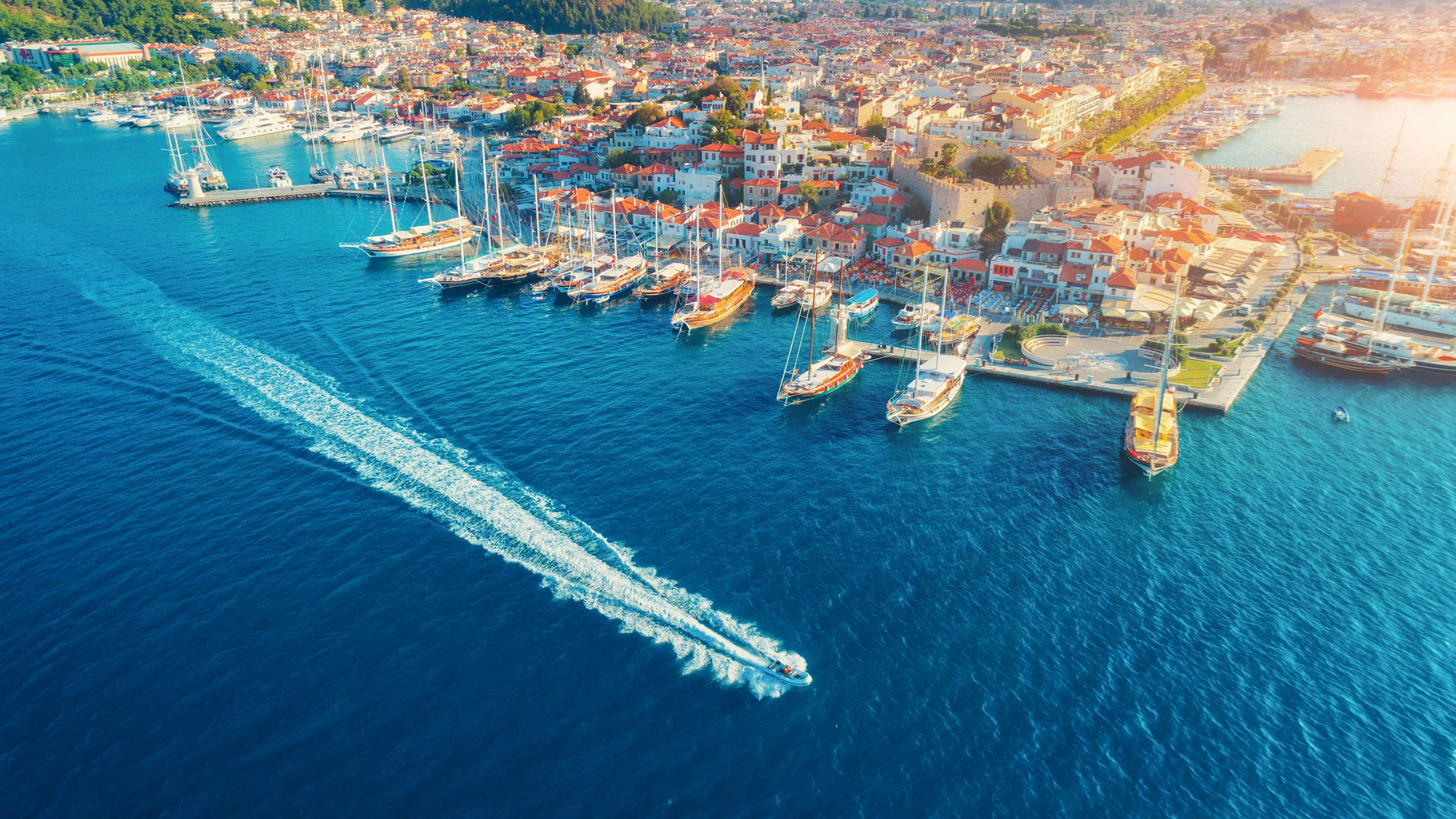 Aerial view of a coastal town with moored yachts and a prominent yacht leaving the harbor.